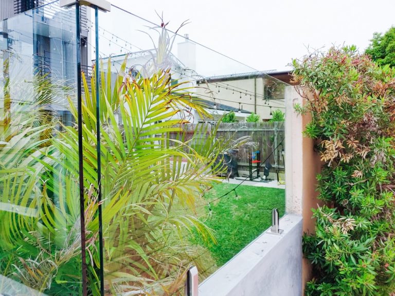 Small section of glass railing mounted to a pony wall surrounding plants and a backyard area