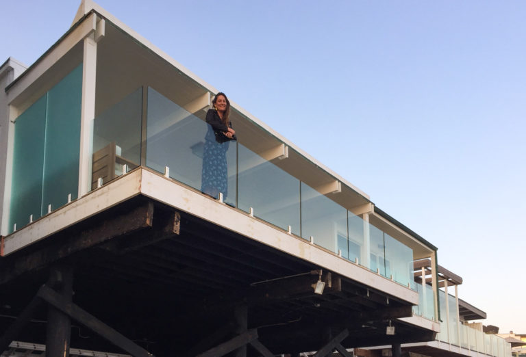 A woman standing on her balcony smiling and leaning against glass railing.